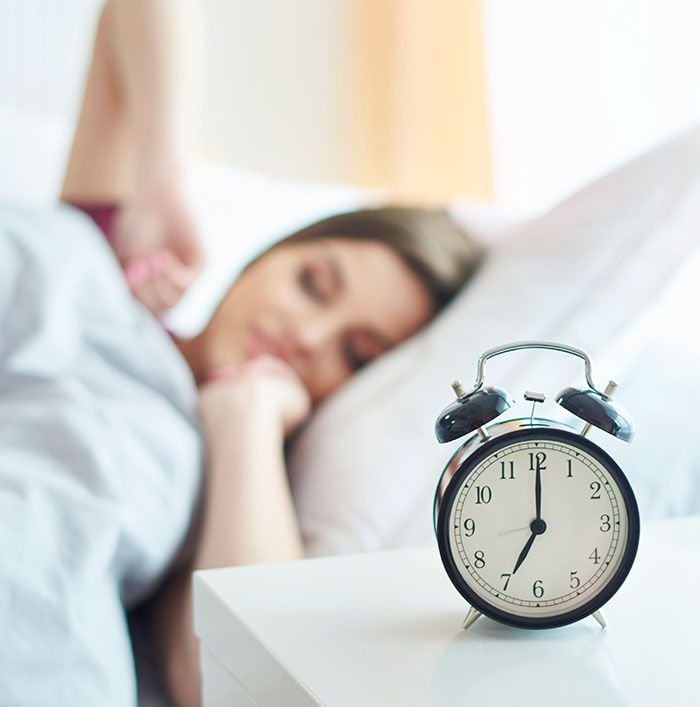 Young woman waking up in bed with soft morning light, stretching with eyes closed and a classic alarm clock on the nightstand — healthy morning routine to boost metabolism and energy naturally.